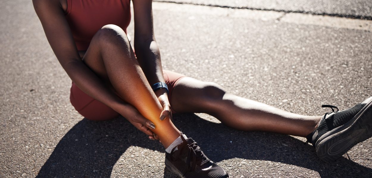 Leg, injury and pain with a sports black woman holding her ankle outdoor during a fitness workout from above. Ankle, sprain or exercise with a female runner or athlete suffering from an injured joint.