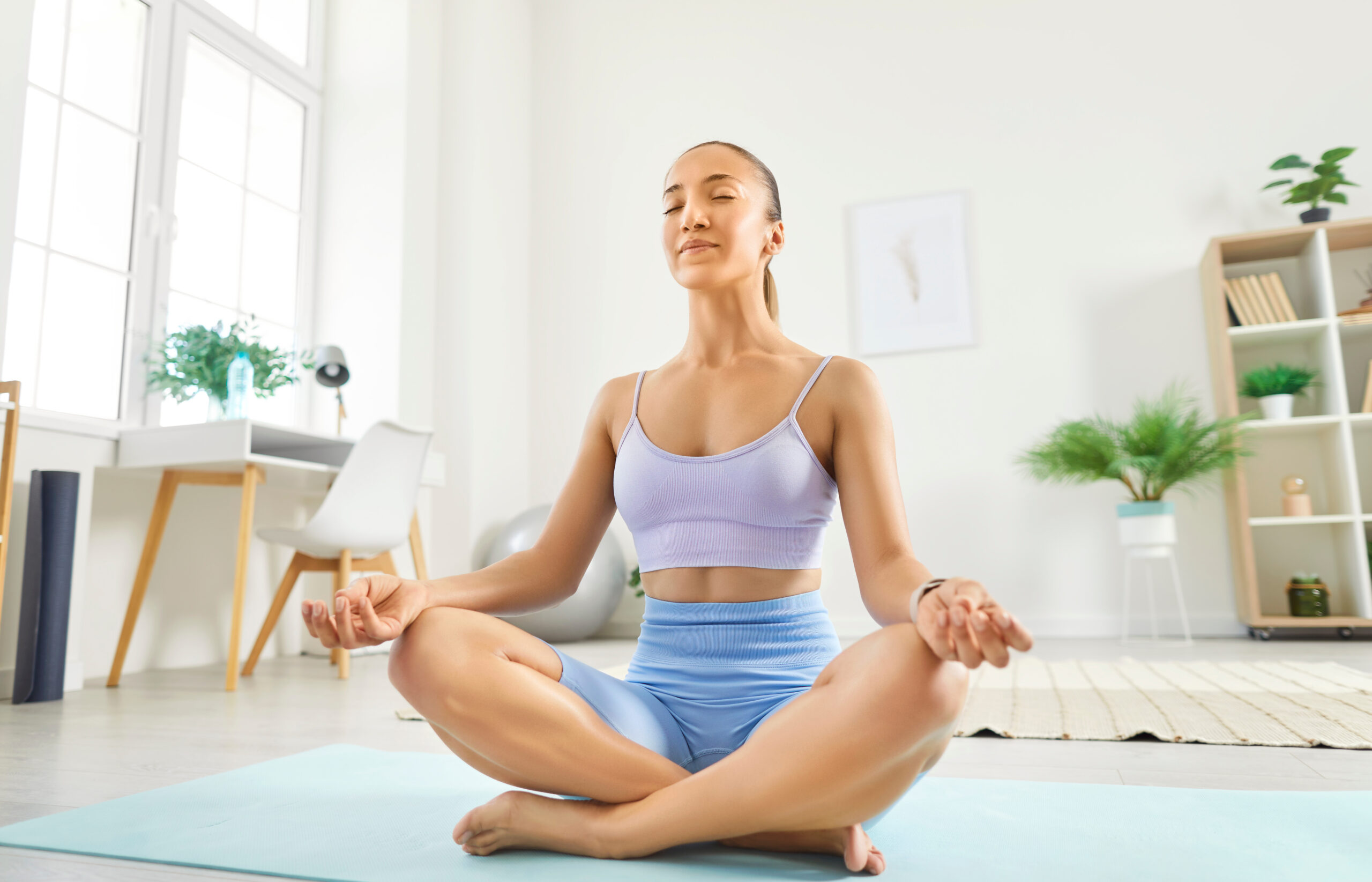 Woman is meditating in the lotus position while practicing yoga at home. Her training session emphasizes relaxation, tranquility, and sport, showcasing a refreshing lifestyle focused on well-being.